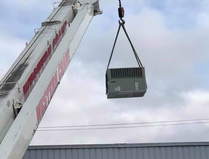 A large commercial rooftop HVAC unit being hoisted into place by a construction crane against a clear blue sky